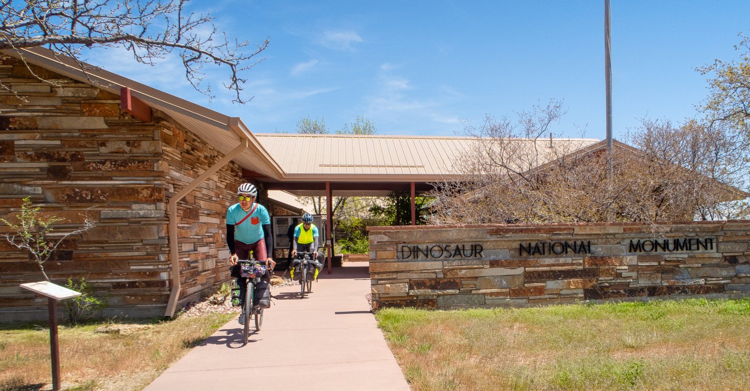 image of fergus and corey riding bikes next to the dinosaur national monument sign